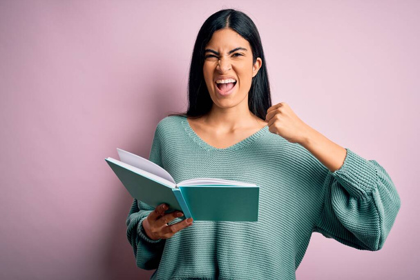 Νεαρή όμορφη Ισπανίδα μαθήτρια που διαβάζει ένα βιβλίο πάνω από ροζ απομονωμένο φόντο ενοχλημένη και απογοητευμένη φωνάζοντας με θυμό, τρελή και φωνάζοντας με σηκωμένο χέρι, έννοια του θυμού - Φωτογραφία, εικόνα