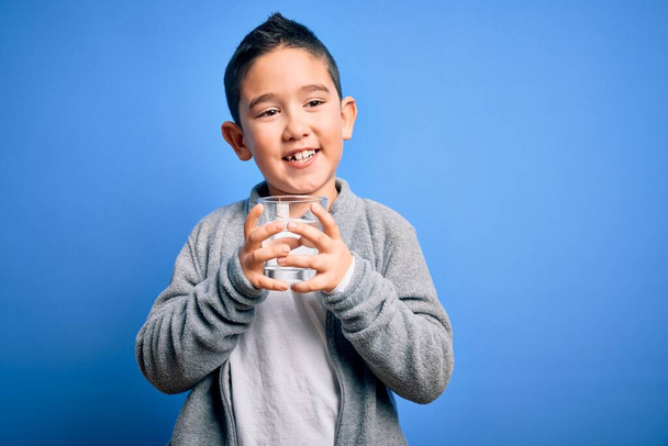 Adorable toddler smiling happy and confident. Standing with smile on face holding glass of healthy water to refreshment over isolated blue background - Photo, Image