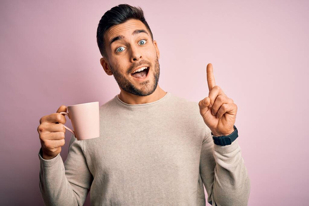 Genç yakışıklı bir adam pembe izole edilmiş arka planda bir fincan sıcak kahve içiyor. Bir fikir ya da soru işareti ile şaşırmış, mutlu bir yüz, bir numara. - Fotoğraf, Görsel