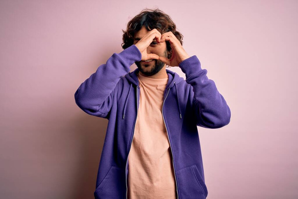 Young handsome sporty man with beard wearing casual sweatshirt over pink background Doing heart shape with hand and fingers smiling looking through sign - Фото, изображение