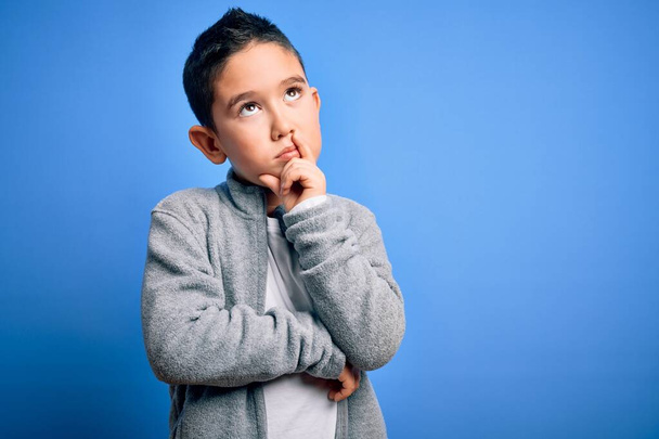 Молодий маленький хлопчик, який носить спортивний светр над синім ізольованим тлом, з рукою на підборідді думає про питання, стомлювальне вираження. Посміхаючись з розсудливим обличчям. Концепція сумніву
. - Фото, зображення