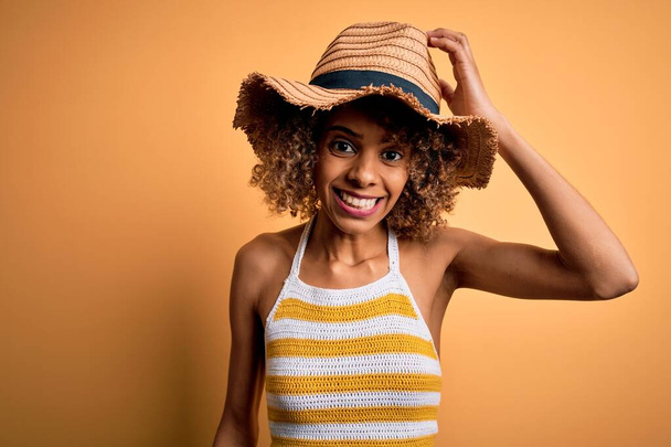 African american tourist woman with curly on vacation wearing summer hat and striped t-shirt confuse and wonder about question. Uncertain with doubt, thinking with hand on head. Pensive concept. - Φωτογραφία, εικόνα