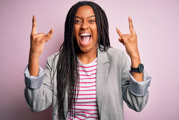 Junge afrikanisch-amerikanische Geschäftsfrau steht vor rosa isoliertem Hintergrund und schreit mit verrücktem Gesichtsausdruck Rock-Symbol mit erhobenen Händen. Musikstar. Konzept für schwere Musik. - Foto, Bild