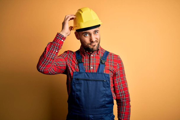 Nuori rakentaja mies yllään rakentaminen yhtenäinen ja turvallisuus kypärä yli keltainen eristetty tausta hämmentää ja ihmetellä kysymys. Epävarma epäilystä, ajatteleva käsi pään päällä. Mietteliäs käsite
. - Valokuva, kuva
