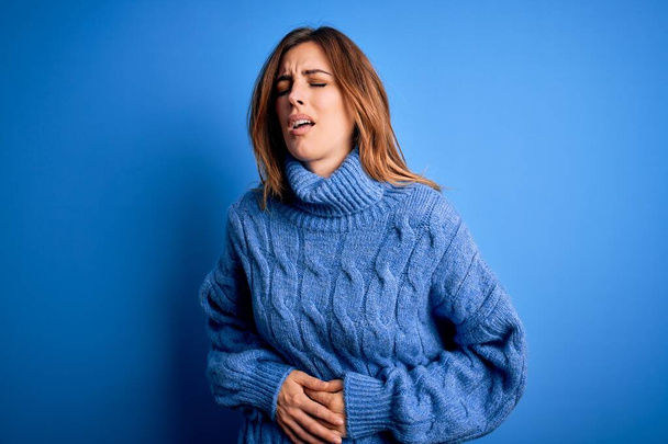 Young beautiful brunette woman wearing casual turtleneck sweater over blue background with hand on stomach because nausea, painful disease feeling unwell. Ache concept. - Photo, Image