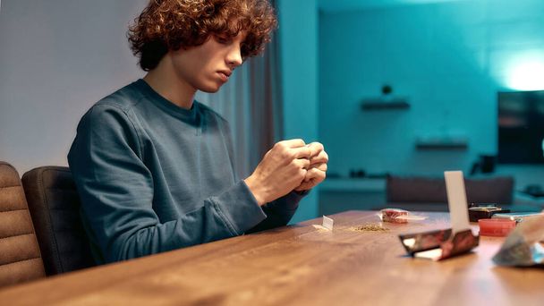 Releasing tension. Young man or teenager making joint, cannabis cigarette sitting at home in the kitchen. Marijuana tools on the table. Cannabis and weed legalization concept - Photo, image