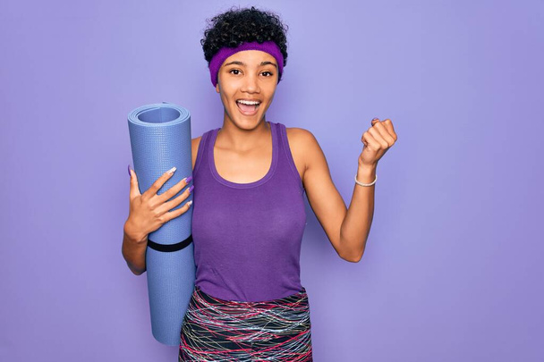 Junge schöne afrikanisch-amerikanische sportliche Frau in Sportbekleidung hält Yogamatte schreiend stolz und feiert Sieg und Erfolg sehr aufgeregt, jubelnde Emotion - Foto, Bild