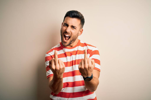 Young handsome man wearing casual striped t-shirt standing over isolated white background Showing middle finger doing fuck you bad expression, provocation and rude attitude. Screaming excited - Zdjęcie, obraz