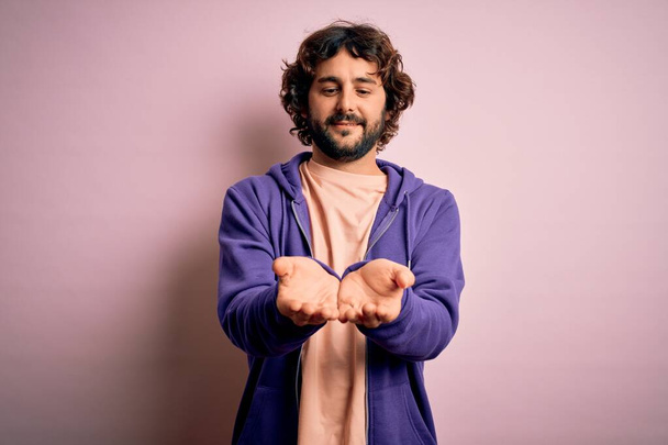 Young handsome sporty man with beard wearing casual sweatshirt over pink background Smiling with hands palms together receiving or giving gesture. Hold and protection - Photo, Image
