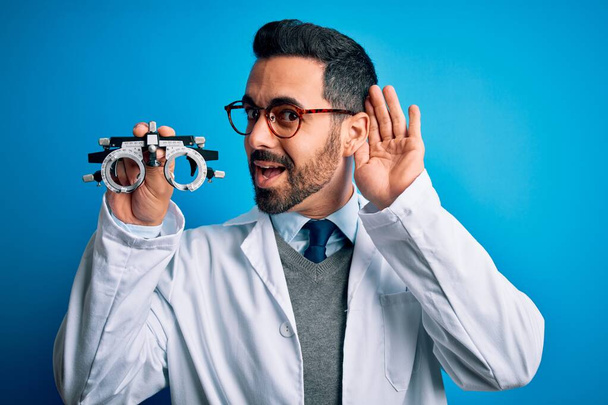 Young handsome optical man with beard holding optometry glasses over blue background smiling with hand over ear listening an hearing to rumor or gossip. Deafness concept. - Photo, Image