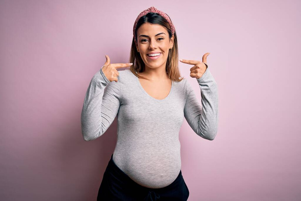 Νεαρή όμορφη μελαχρινή γυναίκα έγκυος αναμένουν μωρό πάνω από απομονωμένο ροζ φόντο χαμογελώντας χαρούμενα δείχνει και δείχνοντας με τα δάχτυλα των δοντιών και του στόματος. Έννοια υγείας οδόντων. - Φωτογραφία, εικόνα