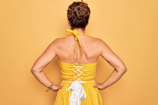 Middle age senior pin up woman wearing 50s style retro dress over yellow background standing backwards looking away with arms on body - Foto, afbeelding