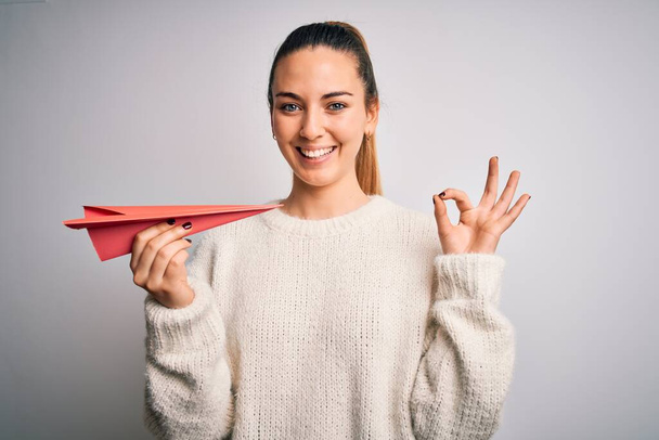 Beautiful blonde woman with blue eyes holding red paper airplane over white background doing ok sign with fingers, excellent symbol - Foto, afbeelding