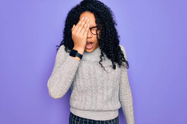 Jeune femme afro-américaine portant un pull décontracté et des lunettes sur fond violet Bâillement fatigué couvrant la moitié du visage, des yeux et de la bouche avec la main. Le visage souffre dans la douleur
. - Photo, image