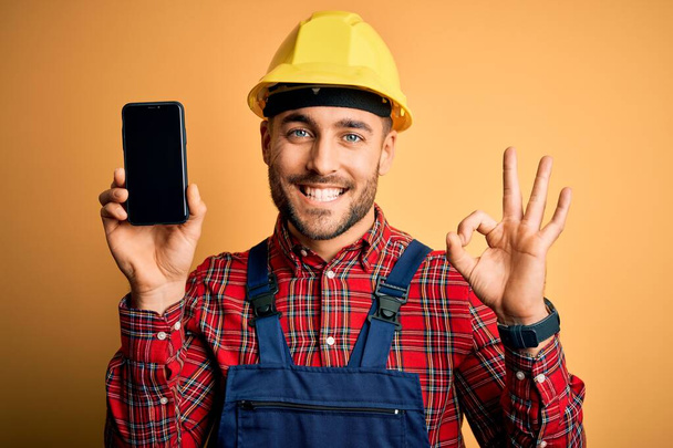 Nuori rakentaja mies yllään turvakypärä osoittaa älypuhelimen näytön keltainen tausta tekee ok merkki sormilla, erinomainen symboli
 - Valokuva, kuva