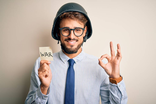 Jonge zakenman met baard die een helm draagt, papier vasthoudend met oorlogsboodschap, goed teken met vingers, uitstekend symbool. - Foto, afbeelding