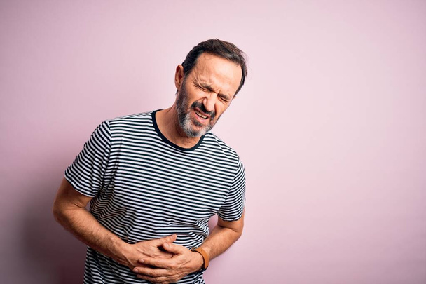 Hombre alzado de mediana edad que usa una camiseta de rayas casuales de pie sobre un fondo rosa aislado con la mano en el estómago porque las náuseas, la enfermedad dolorosa sensación de malestar. Concepto Ache
. - Foto, imagen