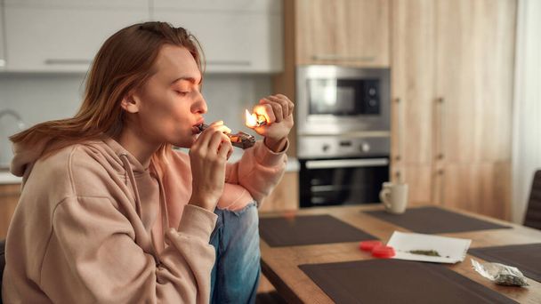 Reducing Stress. Young caucasian woman lighting cannabis in the bowl of metal pipe while sitting in the kitchen. Buds in a plastic bag and red marijuana grinder on the table - Fotó, kép