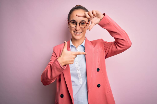 Young beautiful businesswoman wearing jacket and glasses over isolated pink background smiling making frame with hands and fingers with happy face. Creativity and photography concept. - Photo, Image