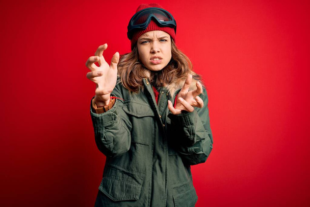 Νεαρό ξανθό κορίτσι που φοράει γυαλιά του σκι και χειμερινό παλτό για τον καιρό του σκι πάνω από το κόκκινο φόντο Φωνάζοντας απογοητευμένοι από την οργή, τα χέρια προσπαθούν να στραγγαλίσουν, φωνάζοντας τρελός - Φωτογραφία, εικόνα