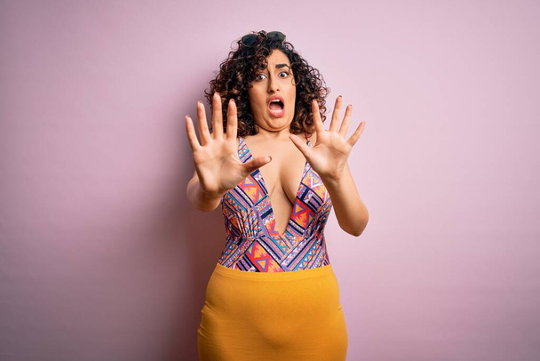 Young beautiful arab woman on vacation wearing swimsuit and sunglasses over pink background afraid and terrified with fear expression stop gesture with hands, shouting in shock. Panic concept. - Photo, Image