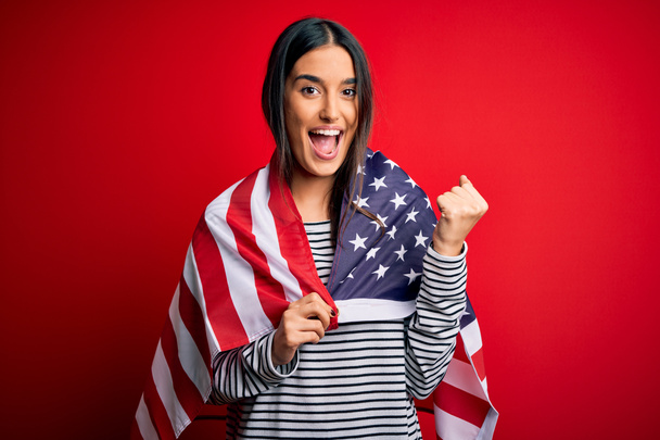 Νεαρή όμορφη μελαχρινή πατριώτισσα γυναίκα φορώντας σημαία των Ηνωμένων Πολιτειών για την ημέρα της ανεξαρτησίας ουρλιάζοντας περήφανη και γιορτάζοντας τη νίκη και την επιτυχία πολύ ενθουσιασμένος, ζητωκραυγάζοντας συναίσθημα - Φωτογραφία, εικόνα