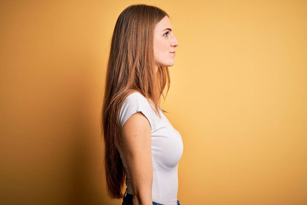 Young beautiful redhead woman wearing casual t-shirt over isolated yellow background looking to side, relax profile pose with natural face and confident smile. - 写真・画像