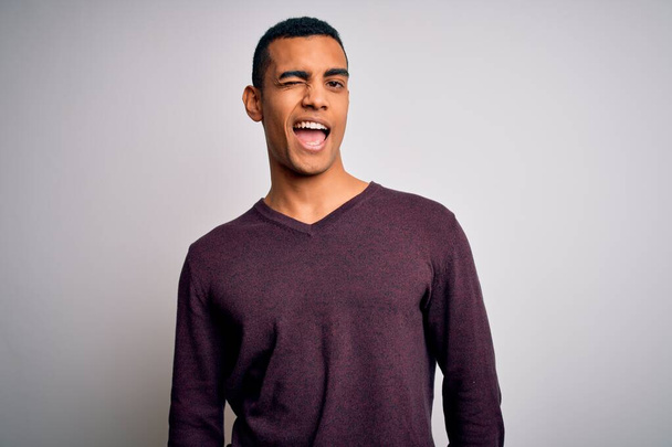 Junger gutaussehender afrikanisch-amerikanischer Mann in lässigem Pullover vor weißem Hintergrund zwinkert in die Kamera mit sexy Ausdruck, fröhlichem und glücklichem Gesicht. - Foto, Bild