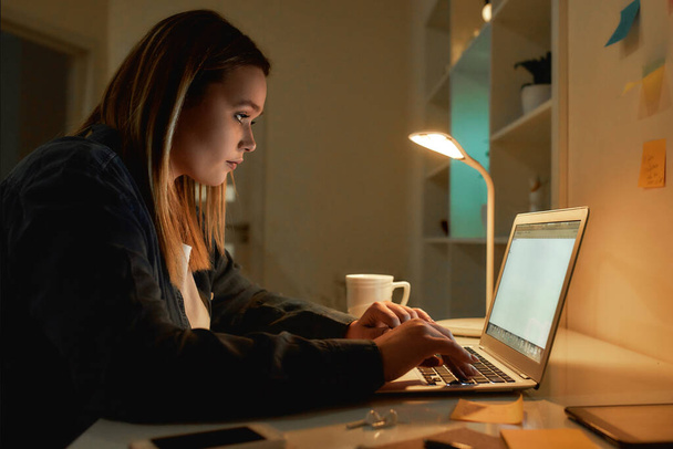 Overworking. Side view of young caucasian woman working from home in dark late night, using laptop and drinking coffee. Sitting at her home office, working remotely - Φωτογραφία, εικόνα