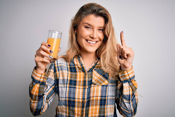 Jovem mulher loira bonita bebendo copo de suco de laranja saudável sobre fundo branco surpreso com uma ideia ou pergunta apontando o dedo com o rosto feliz, número um
 - Foto, Imagem