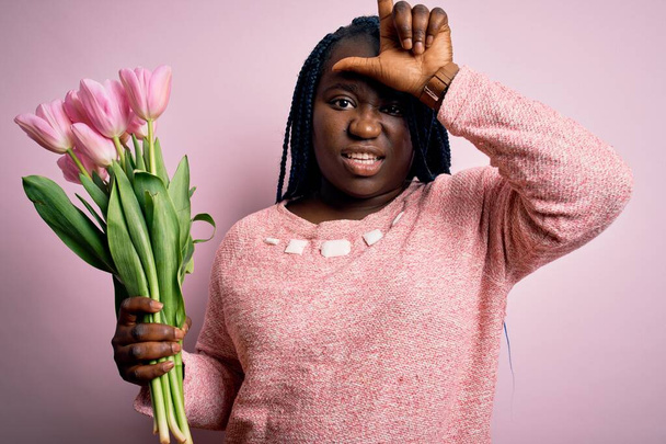 Joven afroamericana mujer de tamaño grande con trenzas sosteniendo ramo de tulipanes rosados flor burlándose de la gente con los dedos en la frente haciendo gesto perdedor burlándose e insultando
. - Foto, imagen