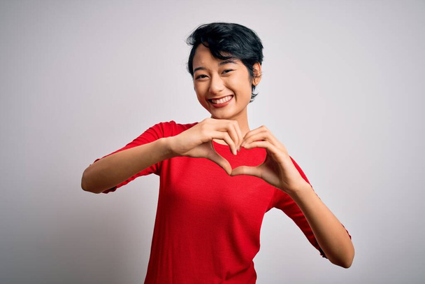 Giovane bella ragazza asiatica indossa casual t-shirt rossa in piedi sopra isolato sfondo bianco sorridente in amore facendo forma di simbolo del cuore con le mani. Concetto romantico
. - Foto, immagini