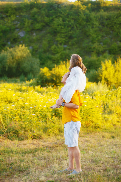 Ojciec trzyma na ramionach jego córeczkę w białej sukience spoglądającą daleko. - Zdjęcie, obraz