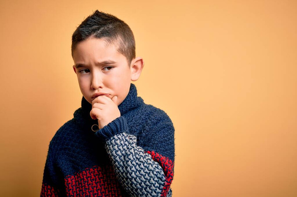 若い男の子の冬のセーターを黄色の隔離された背景に手であごの質問について考える上で身に着けている、集中的な表現。思慮深い顔で微笑んだ。疑いの概念. - 写真・画像