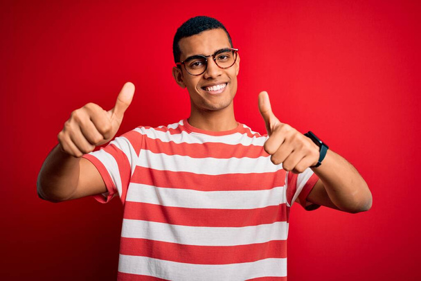 若いハンサムなアフリカ系アメリカ人の男性は、カジュアルなストライプのTシャツと眼鏡を着用し、手で積極的なジェスチャーを行うことを承認し、親指を笑顔と成功のために幸せ。勝者のジェスチャー. - 写真・画像