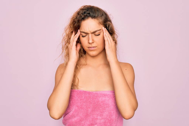 Schöne blonde Frau mit blauen Augen, die Handtuchdusche nach dem Bad über rosa Hintergrund mit der Hand auf dem Kopf für Kopfschmerzen wegen Stress trägt. Migräne. - Foto, Bild