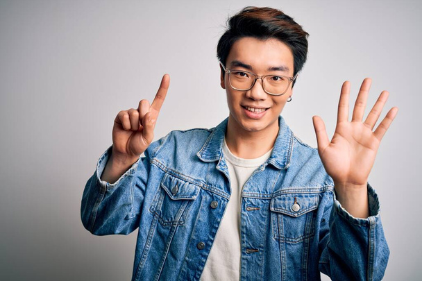 Jeune bel homme chinois portant une veste en denim et des lunettes sur fond blanc montrant et pointant vers le haut avec les doigts numéro six tout en souriant confiant et heureux
. - Photo, image