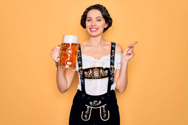 Krásná německá žena s modrýma očima na sobě tradiční octoberfest šaty pití sklenice piva velmi šťastný ukazující rukou a prstem na stranu - Fotografie, Obrázek