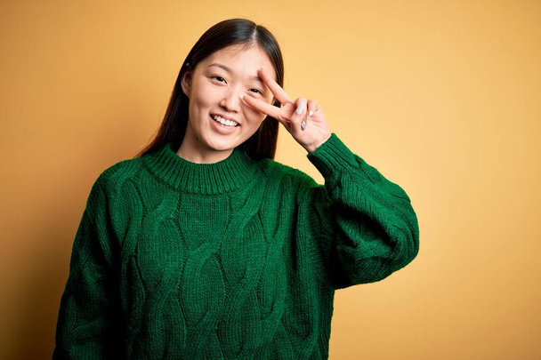 Młoda piękna azjatycka kobieta w zielonym zimowym swetrze na żółtym odizolowanym tle Robi symbol pokoju z palcami na twarzy, uśmiechnięta radosna pokazując zwycięstwo - Zdjęcie, obraz