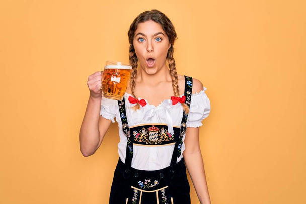Gyönyörű szőke német nő kék szemekkel visel oktoberfest ruha ivás üveg sört megijedt a sokk egy meglepetés arc, fél, és izgatott a félelem kifejezés - Fotó, kép