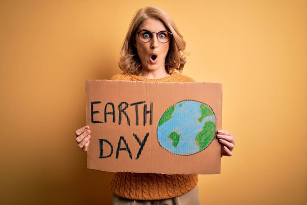 Middle age blonde woman asking for the enviroment holding banner with earth day message scared in shock with a surprise face, afraid and excited with fear expression - Photo, image