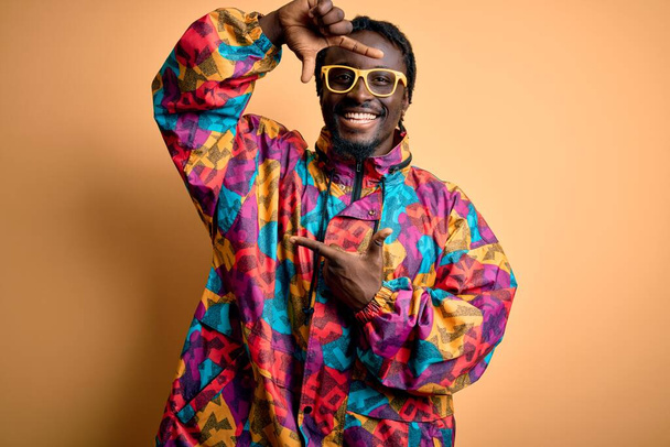 Knappe Afro-Amerikaanse man draagt kleurrijke jas en bril over gele achtergrond glimlachende maken frame met handen en vingers met gelukkig gezicht. Creativiteit en fotografie concept. - Foto, afbeelding