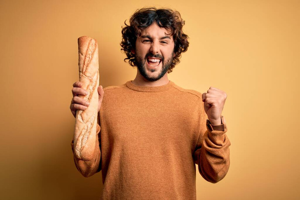 Kıvırcık saçlı ve sakallı genç yakışıklı adam ev yapımı sağlıklı ekmek tutuyor gururla bağırıyor ve zaferi ve başarıyı kutluyor. - Fotoğraf, Görsel