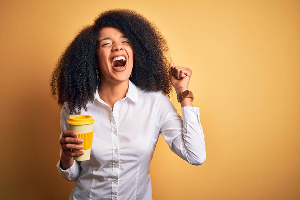 Νεαρή αφροαμερικανίδα επιχειρηματίας με αφρο μαλλιά πίνοντας καφέ από το take away cup ουρλιάζοντας περήφανη και γιορτάζοντας τη νίκη και την επιτυχία πολύ ενθουσιασμένος, ζητωκραυγάζοντας συγκίνηση - Φωτογραφία, εικόνα