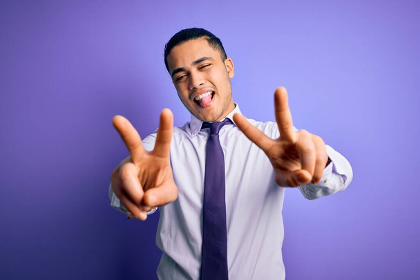 Молодий бізнесмен, одягнений в елегантну краватку, стоїть над ізольованим фіолетовим фоном, усміхаючись язиком, показуючи пальці обох рук, які роблять знак перемоги. Номер два. - Фото, зображення