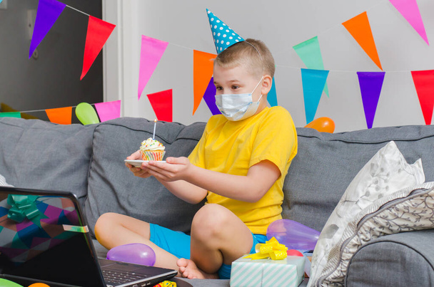 Happy boy in medicine face mask with holiday cupcake celebrates birthday by video call to laptop. Quarantine birthday online in isolation. Virtual event keeping social distance. - Photo, Image