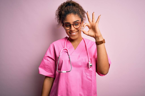 Африканська медсестра з Америки, одягнена в медичну форму і стетоскоп над рожевим фоном, посміхається позитивно, роблячи знак з рукою і пальцями. Успішний вираз. - Фото, зображення