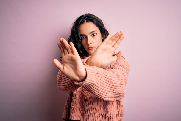 Young beautiful woman with curly hair wearing casual sweater over isolated pink background Rejection expression crossing arms and palms doing negative sign, angry face - Photo, Image