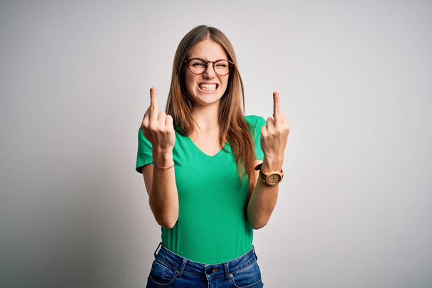 Young beautiful redhead woman wearing casual green t-shirt and glasses over white background Showing middle finger doing fuck you bad expression, provocation and rude attitude. Screaming excited - Foto, Bild