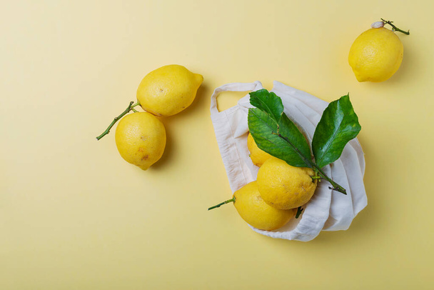 Concept of a biological healthy food and zero waste. Fresh biological lemons with green leaves in a cotton shopping bag on a yellow background. Top view image with a copy space - Zdjęcie, obraz
